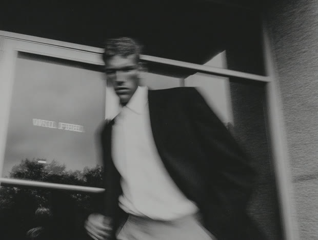 Black-and-white shot of a man carrying a pole past a building