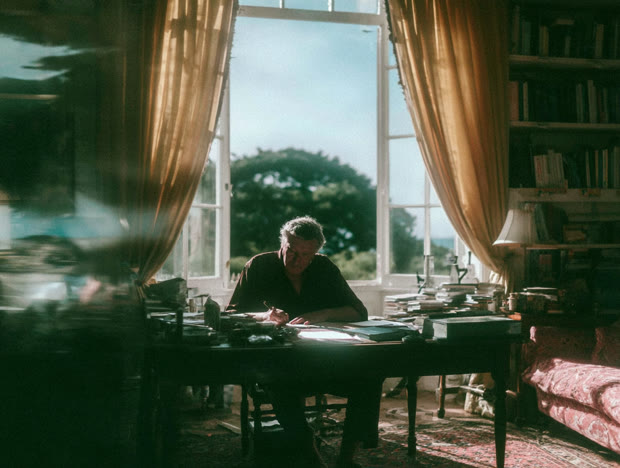 Writer at a desk between tall curtained windows